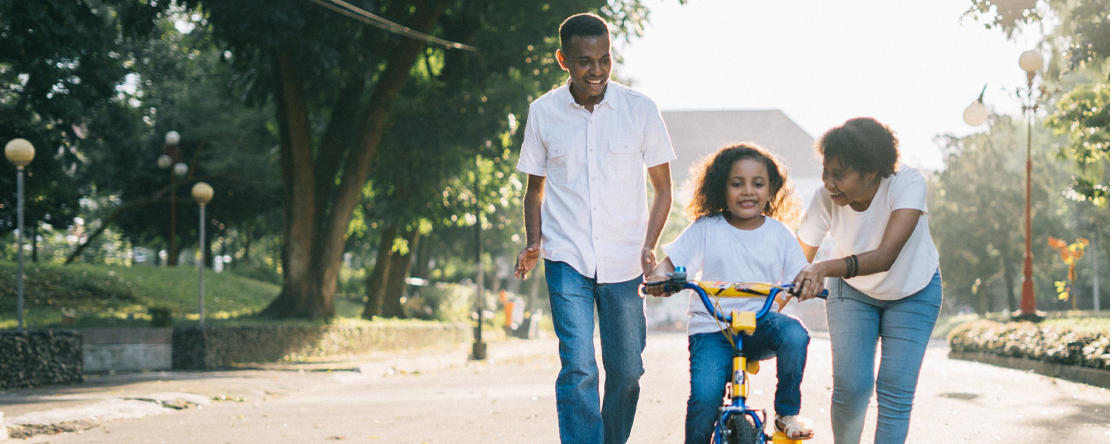 family teaching boy how to ride a bike in neighborhood