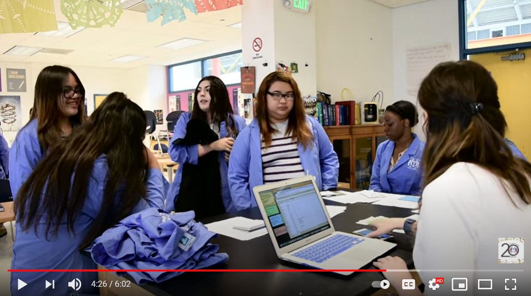 Screenshot from FACES video: Students in blue labcoats, around a table