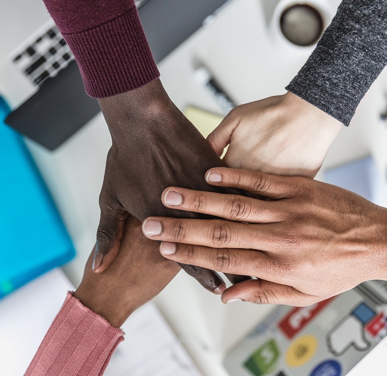 Four people's hands placed together, over computers and paper