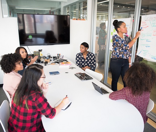 Six women standing and sitting inside a room, having a meeting