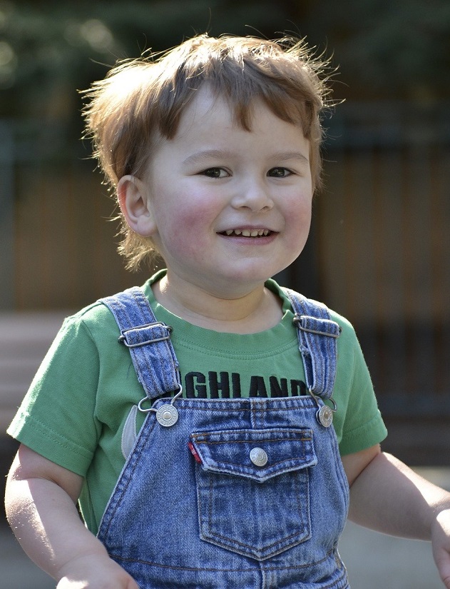 Child in overalls and green t-shirt, smiling at camera