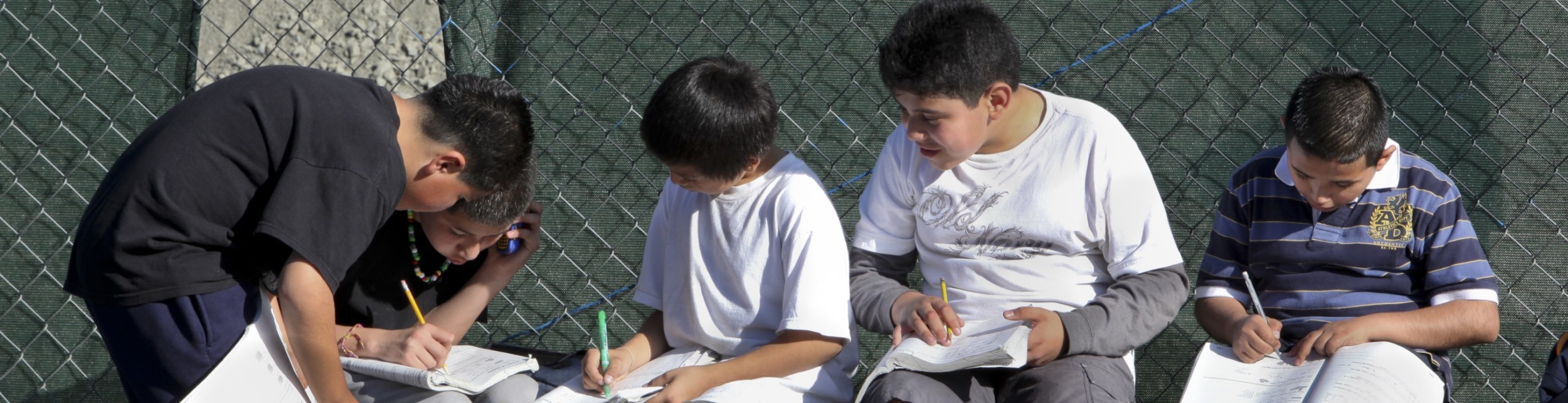 four boys with their backs to a chain-link fence, doing schoolwork together and sharing notes