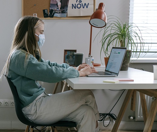 A woman sits in front of a computer wearing a COVID mask