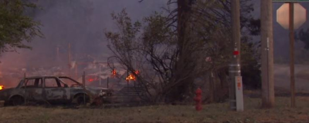 a burned car sits in a smoky haze in front of a burning background