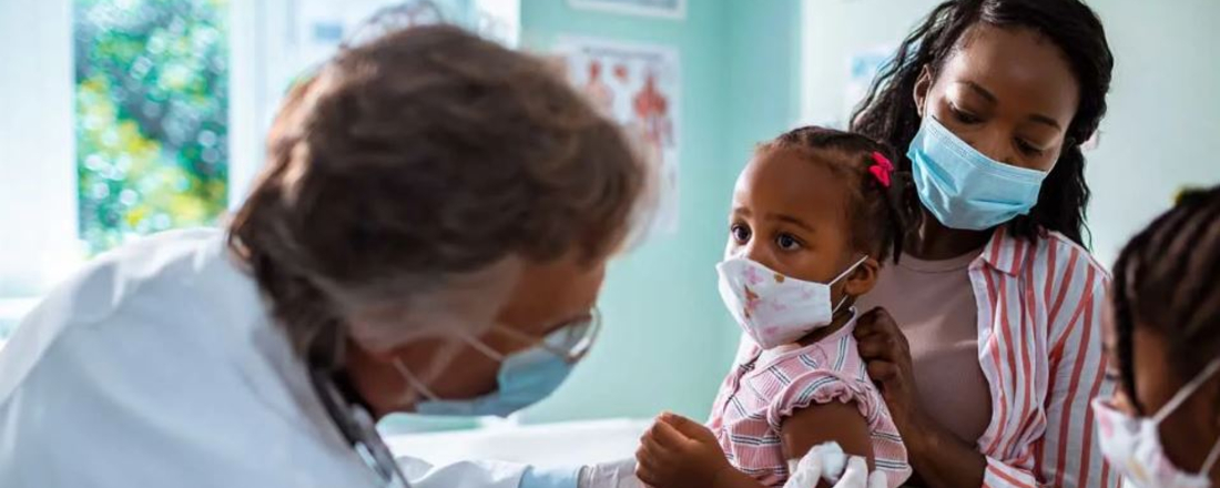 a Mom holds her small child, both in masks, while a doctor gives the child a shot