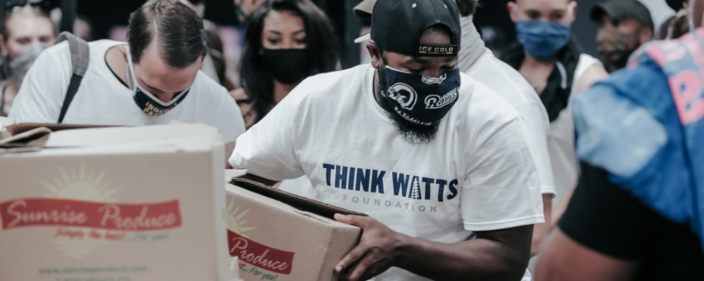 man with a mask and a backward lakers cap unloading food boxes from a stack. his shirt says think watts foundation