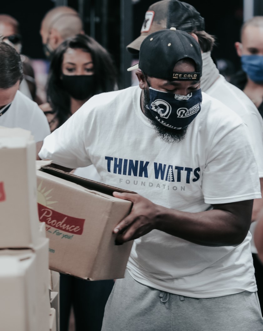 man with a mask and a backward lakers cap unloading food boxes from a stack. his shirt says think watts foundation