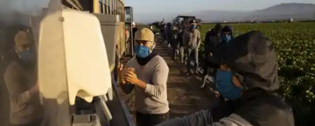 a line of farmworkers in masks waiting to wash their hands alongside a field