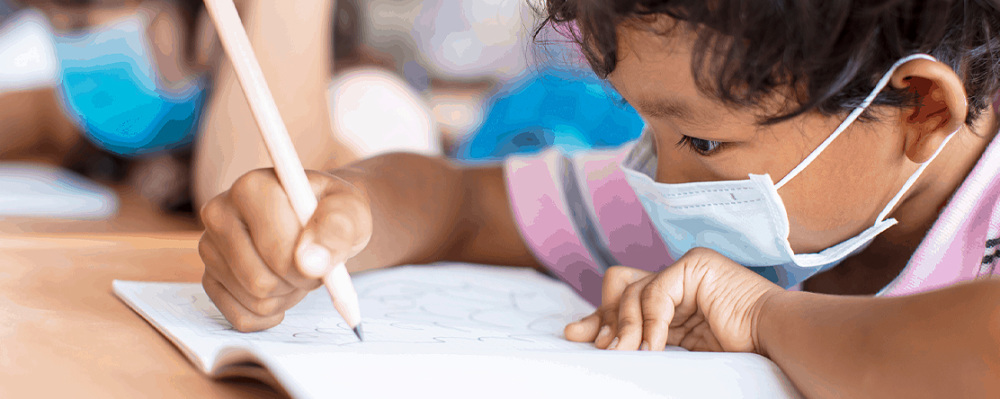 child wearing masks drawing on a notebook in class