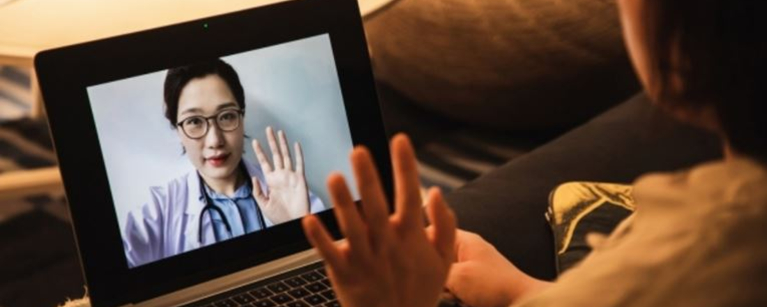 a laptop screen shows a doctor with her hand up while a patient looking on puts a hand up