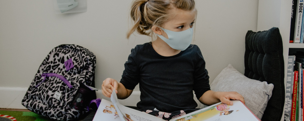 a young child in a mask sitting cross-legged on the floor reading a large kids' book