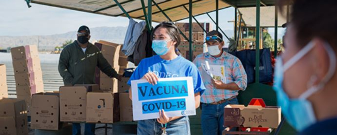 a vaccination clinic for farmworkers with a staff person holding a sign reading "Vacuna COVID-19". Courtesy California Health Line / @heidi_demarco;