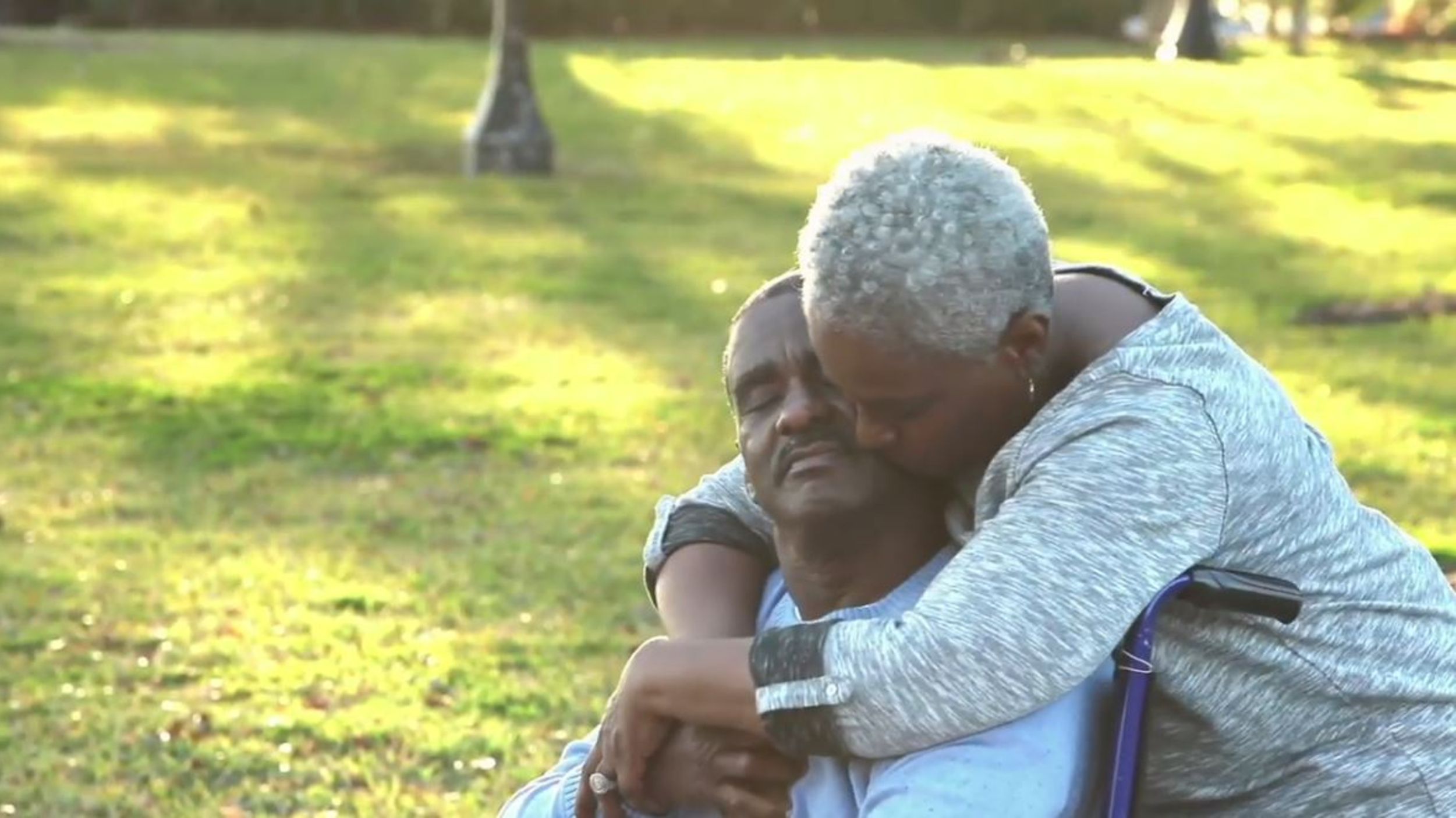 an older African-American couple, the woman hugging the man in a wheelchair from behind