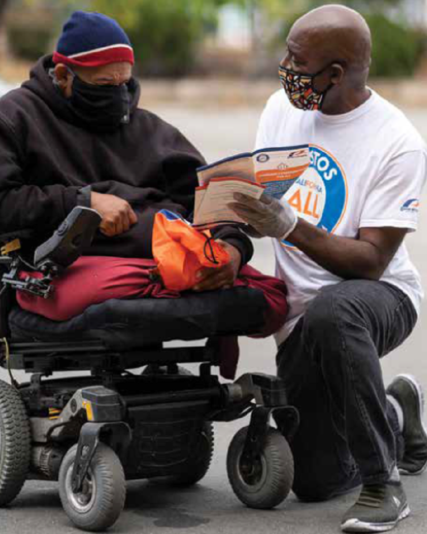 Two men speaking, one is in a wheelchair, the other is kneeling on one knee in front of him showing him a pamphlet.