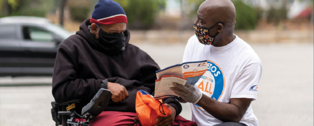 Two men speaking, one is in a wheelchair, the other is kneeling on one knee in front of him showing him a pamphlet.