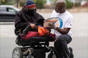 A man in a wheelchair receiving Listos California materials from another man, kneeling beside him. Both are wearing masks