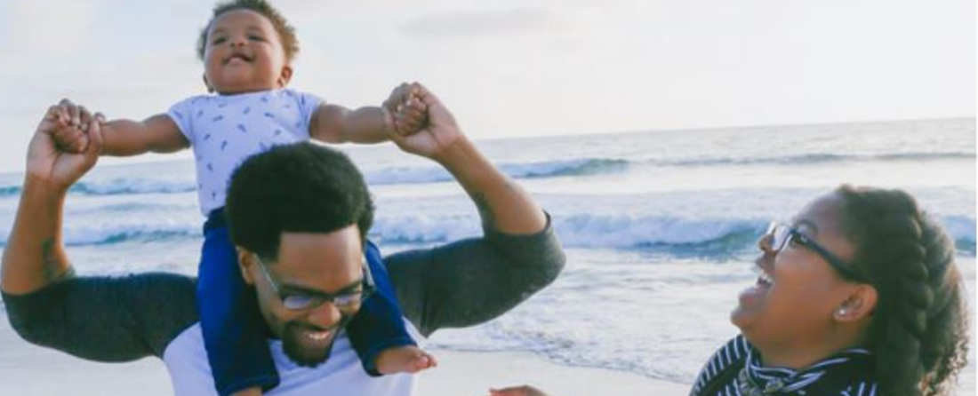 an African American dad and mom at the beach, the dad with a young child on his shoulders