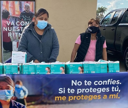 Two women stand in front of a vaccine distribution center at a table with pamphlets in Spanish and English