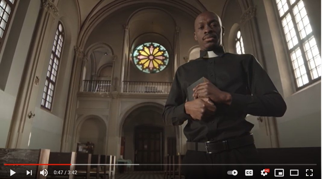 a black pastor standing in a church sanctuary, holding a bible and looking down at the camera