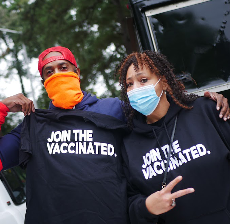 two volunteers wearing masks and tshirts that say "JOIN THE VACCINATED"