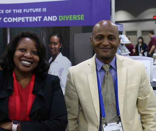 Two people, smiling and looking to camera, inside a conference hall. Behind them, a sign reads "The workforce of the future is competent and diverse"