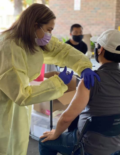 a woman health care worker in a medical gown and mask gives a seated man a vaccination