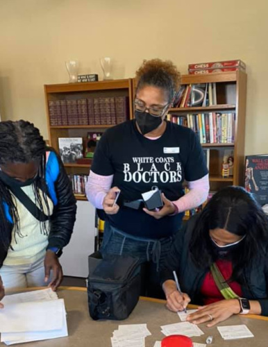 Three African American women in medical masks working together around a table, one in the center with a t-short reading "white coats, Black doctors"