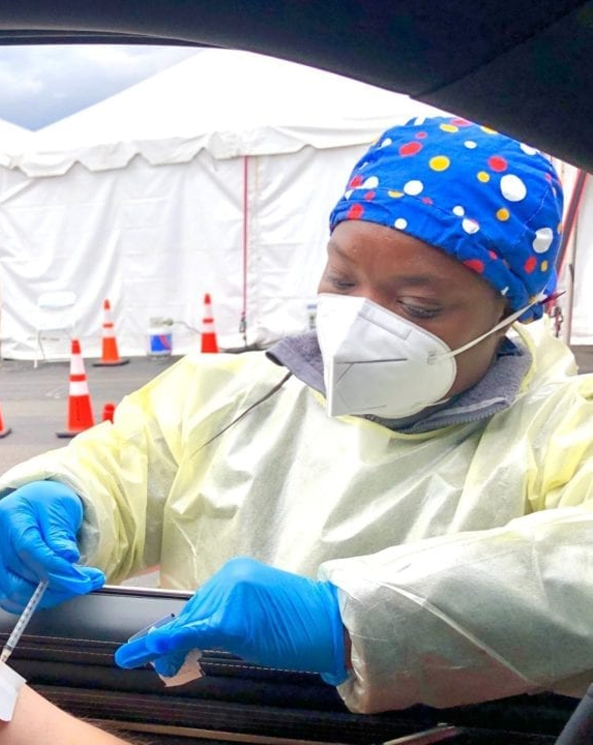 a woman in a medical mask, gown and gloves giving a vaccine through a car window