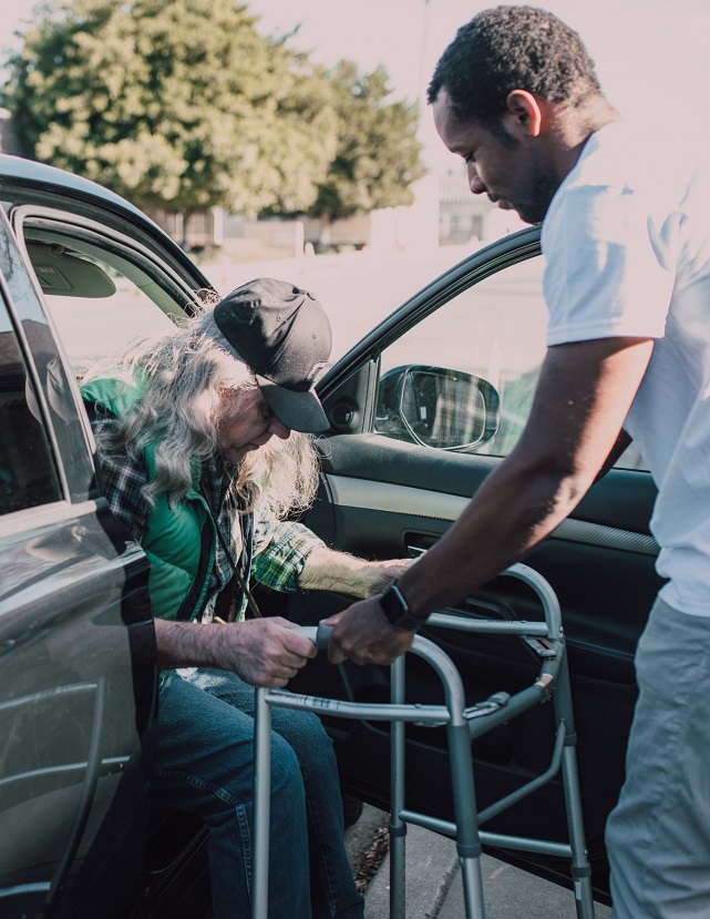 Older adult using a walker to get out of a car, with assistance from a younger man