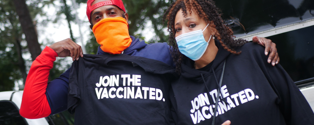 Two people wearing masks and shirts that say "join the vaccinated"