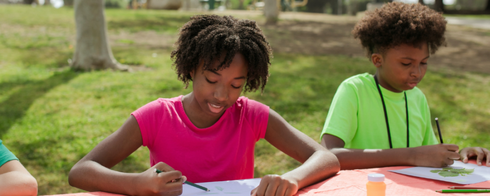 An African American girl sitting next to an African American boy at the park and both are coloring