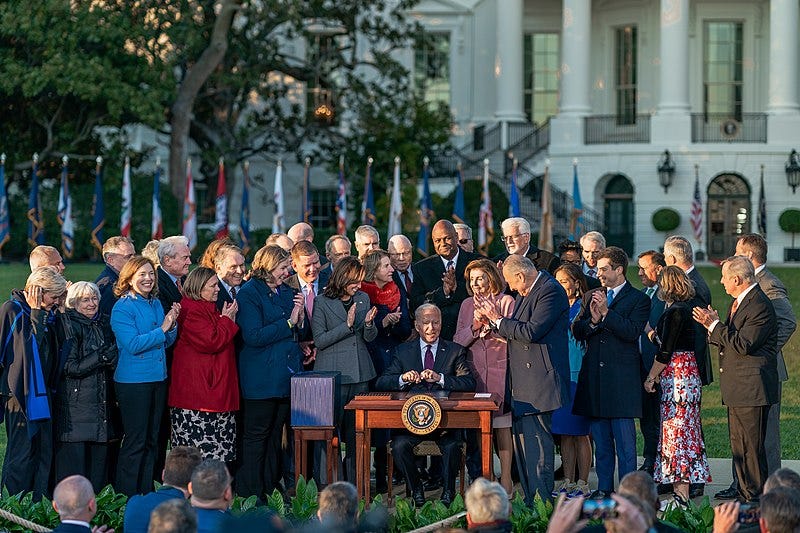 A group of people applaud as they stand around President Biden, sitting outside the White House at a desk with the Great Seal of the U.S.