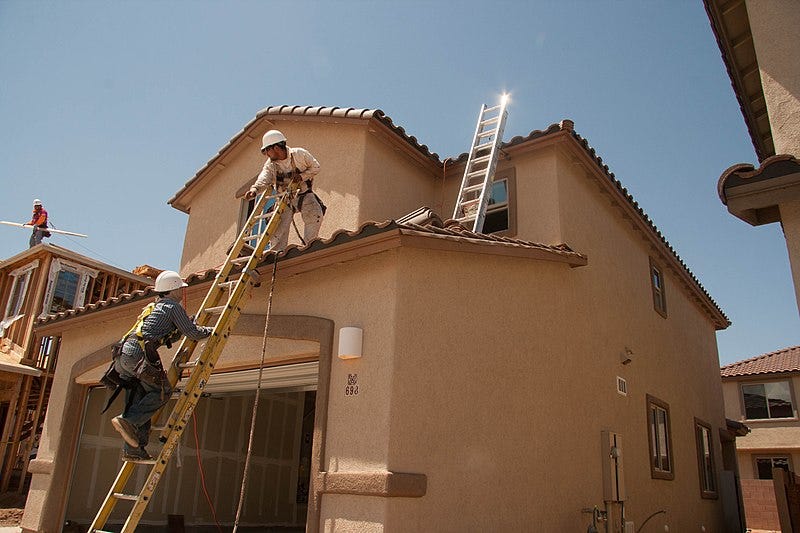 Two men working on a house, one man is climbing a ladder to the roof where the other man stands holding the top of the ladder.