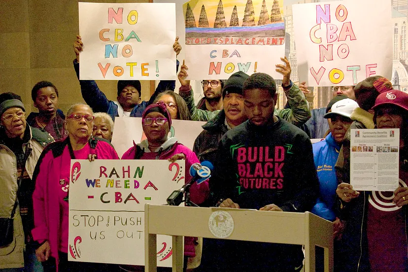 A young Black man stands at a podium giving remarks as a group of people stand behind him, several holding homemade signs protesting a proposed housing vote.