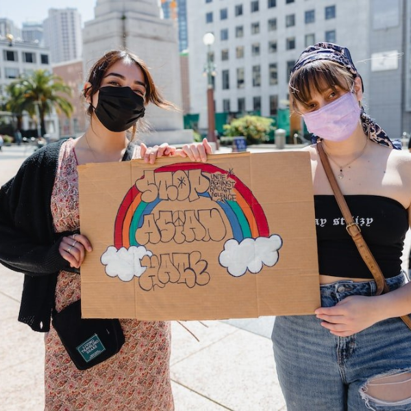 Two people in masks are standing outside holding a sign with a rainbow that reads "Stop Asian Hate."