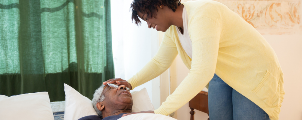 older male laying in bed with caregiver by his side