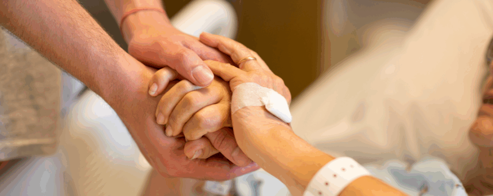 two people hold hands as one lays wearing a hospital intake bracelet in a bed