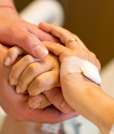 two people hold hands as one lays wearing a hospital intake bracelet in a bed