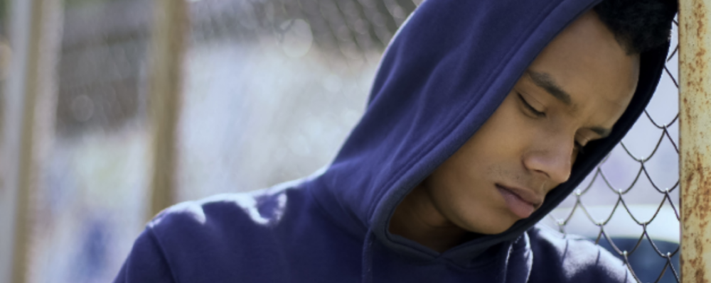 person in a hoodie leaning their head against a chain link fence