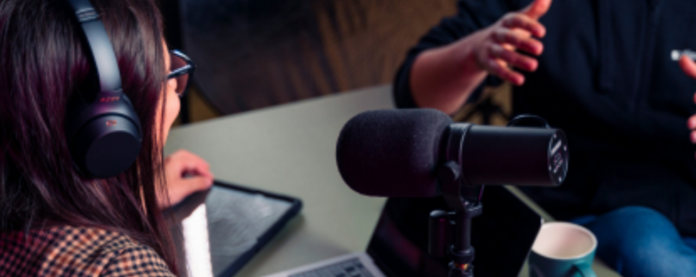 a person wearing headphones sits in front of a microphone across from another person, talking