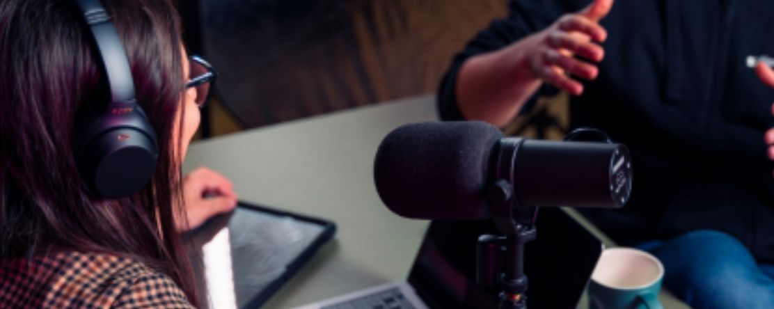 a person wearing headphones sits in front of a microphone across from another person, talking